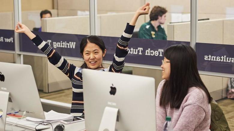 two 学生 sitting together at computer stations, student at right smiles with arms raised joyfully